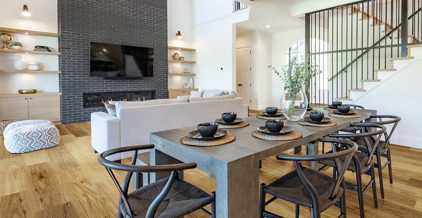 Open living plan with warm-toned hybrid flooring, featuring a cream sofa, charcoal birck feature wall, and dark timber dining table with black woven dining chairs .