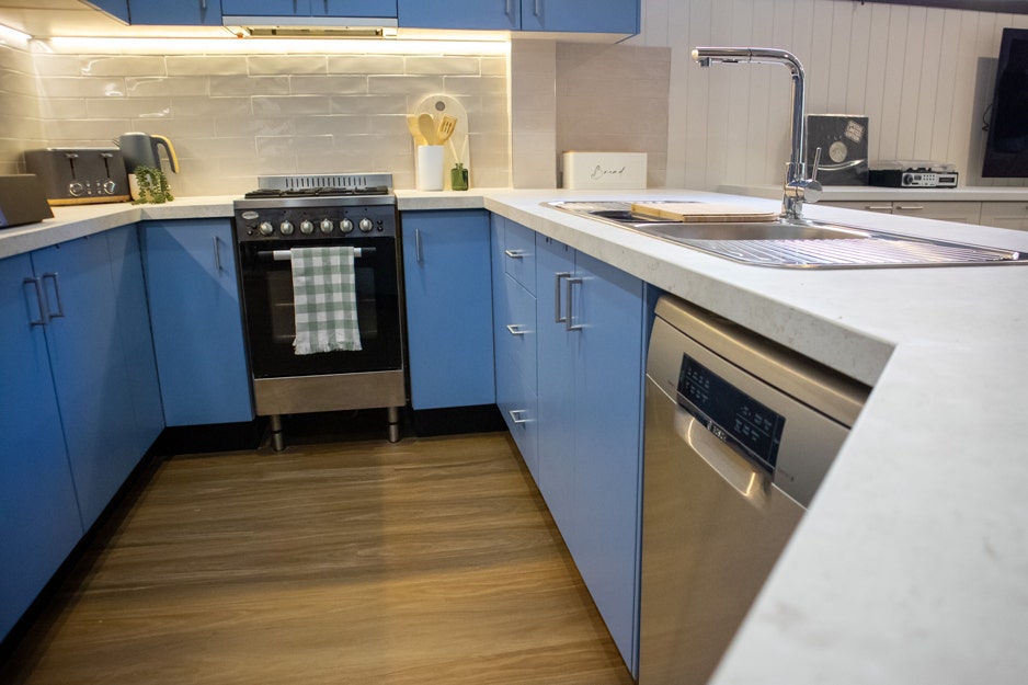 Modern kitchen with stainless steel appliances (sink, dishwasher, stove, oven). White cabinets, light hardwood floors, large window.